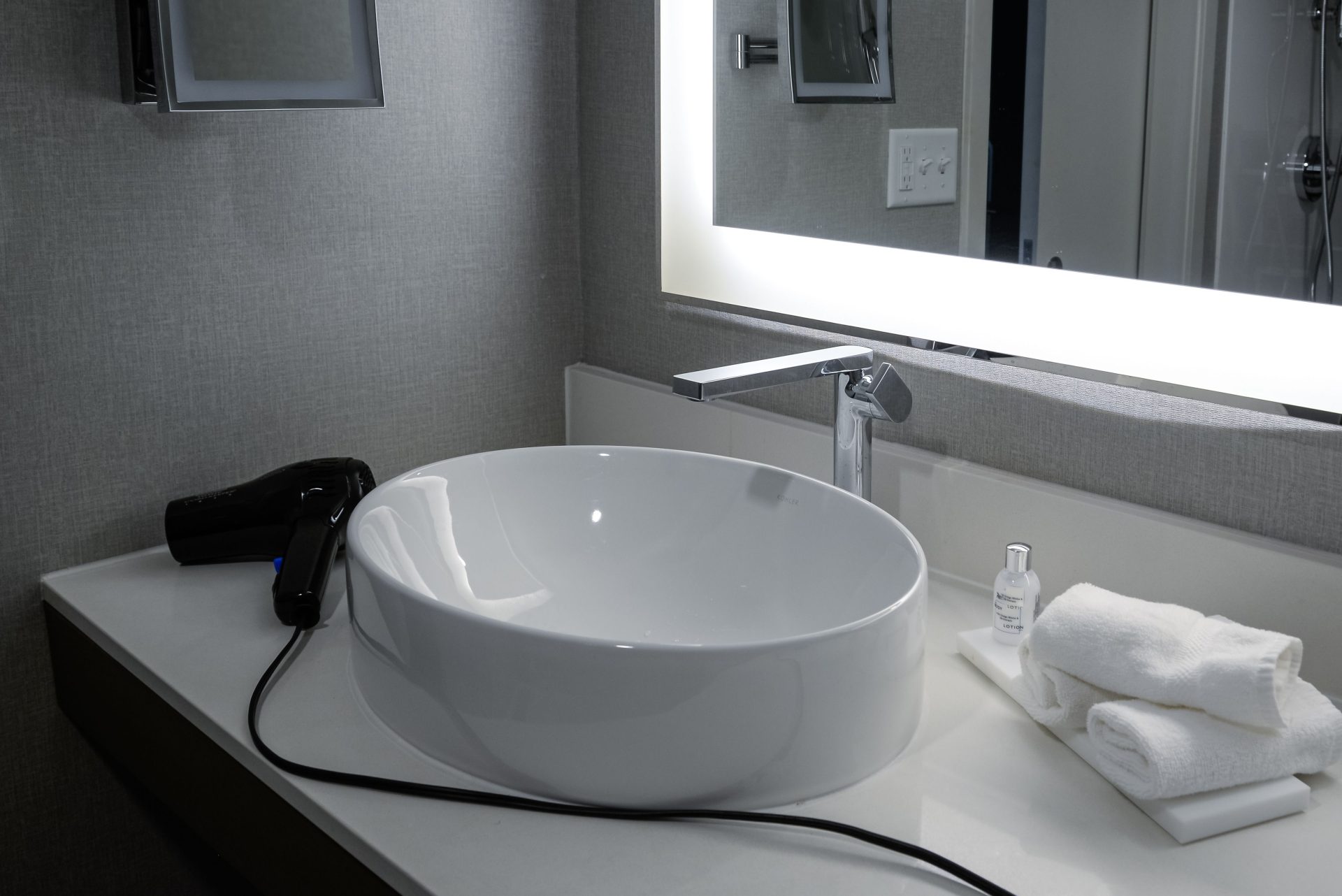 Elegante tocador de baño con lavabo de bol, espejo iluminado y secador de pelo en un entorno hotelero.